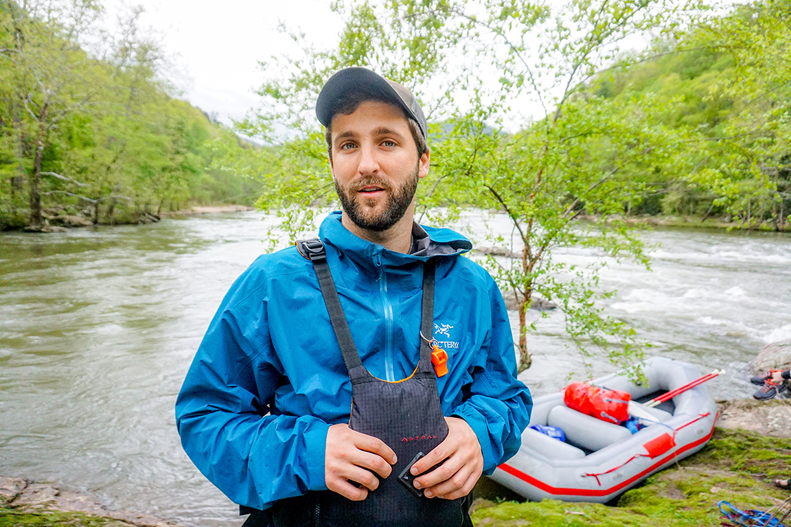 Travis Bordley standing in front of raft and river surrounded by mountains