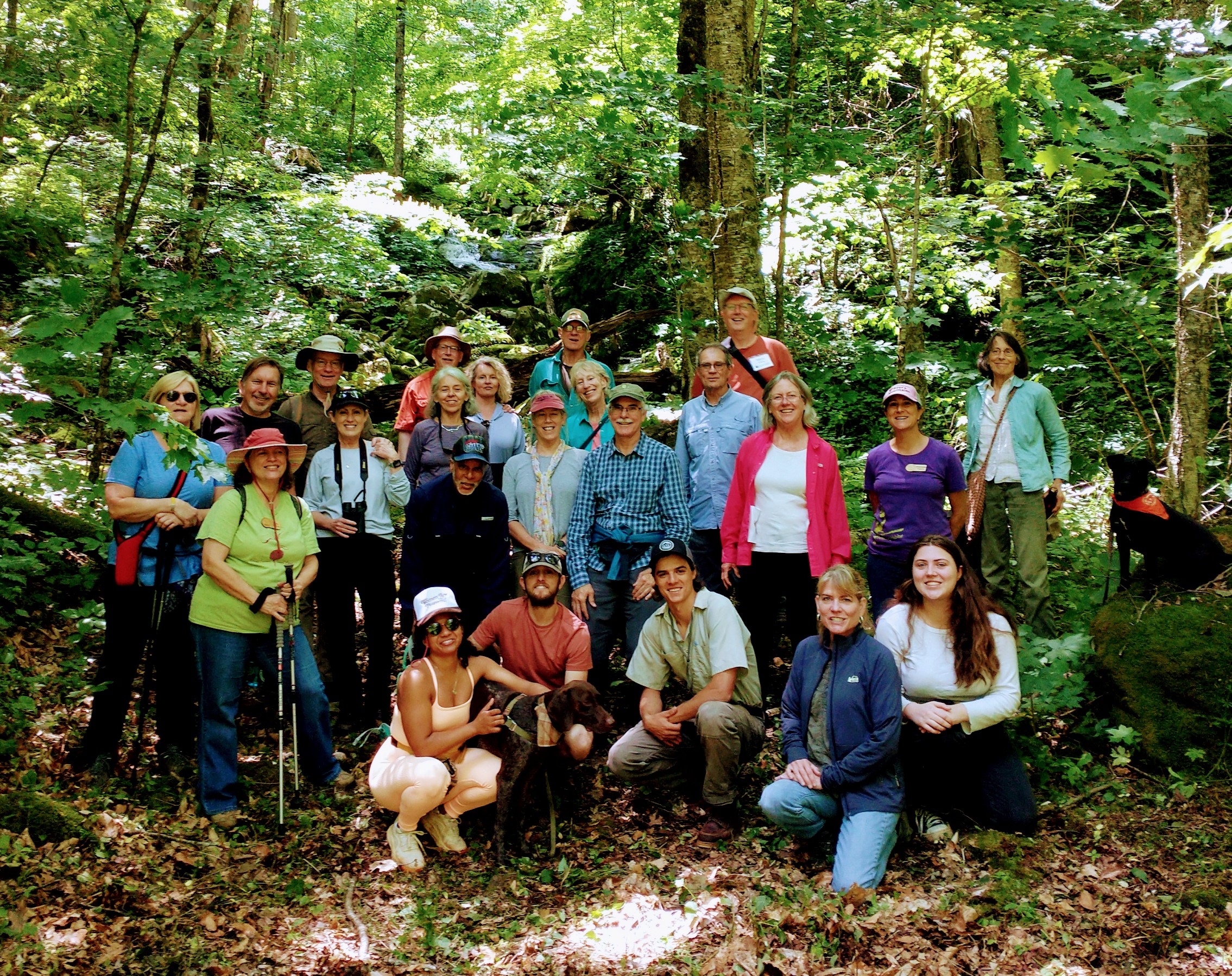 Group of about 25 hikers and one dog posed in front of waterfall and wooded forest trail