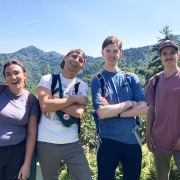 Group of four young adults smiling, some with arms crossed, with mountain ridgeline in background