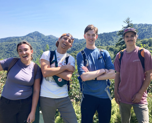 Group of four young adults smiling, some with arms crossed, with mountain ridgeline in background