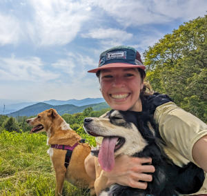 A young women in hat, tan shirt, and backpack hugs two dogs in open area surrounded by forest and distant mountain views.