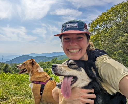PXL_20250611_210947198.MP (1) A young women in hat, tan shirt, and backpack hugs two dogs in open area surrounded by forest and distant mountain views.