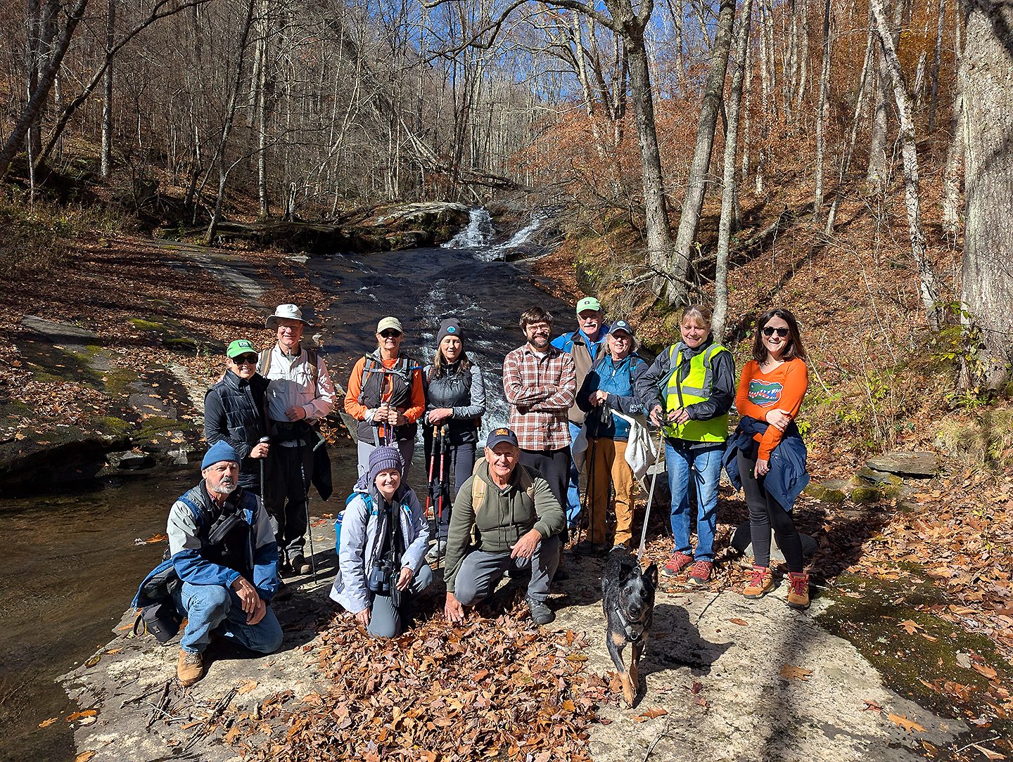 Group of 12 people and one dog posing in front of stream cascade, surrounded by trees and brown and orange fall foliage, and lots of fallen leaves on ground