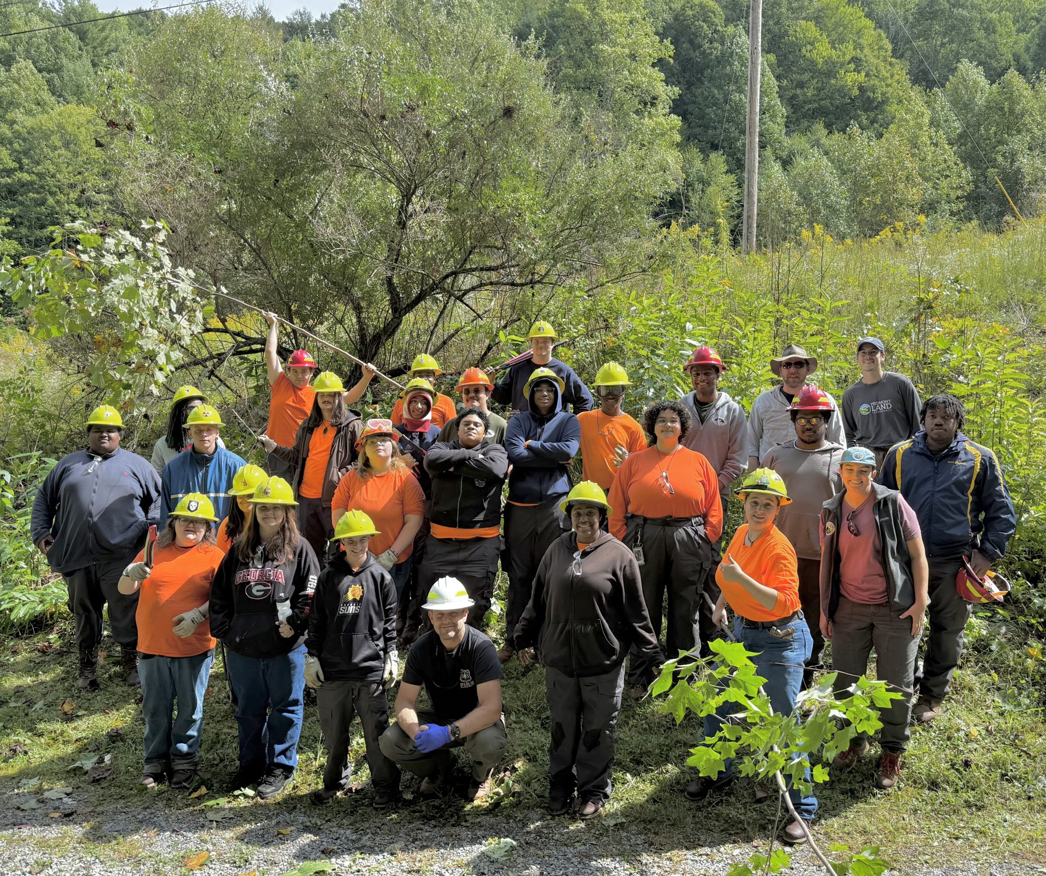 Group of people in bright orange, some with yellow hard hats, standing and kneeling in open area with forest behind them, facing camera
